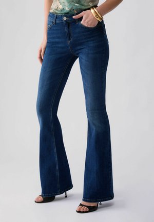 Kvinde iført mørkeblå vidde jeans, grøn blomsterprint top stoppet ind, sorte åbne højhælede sko og gyldne armbånd på håndleddet, står med den ene hånd i lommen.