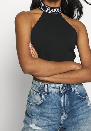 Žena se zkříženýma rukama, oblečená v černém halter topu s logem "KANI" a vysokým pasem roztrhaných modrých džínů, na jednobarevném pozadí.