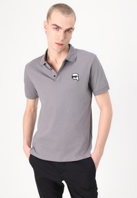 Pelēks kokvilnas polo krekls ar trīs pogu aizdari un melnbaltu grafisku logo uz krūtīm. Īsas piedurknes un klasiskā piegriezuma.
