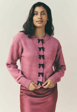 Rosa stickad kofta med rund hals, långa ärmar och svarta rosett-accenter framtill, kombinerad med en glansig rosa satinkjol.