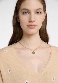 Beige kötött pulóver V-nyakkal, fehér virágmintával hímezve. Finom arany nyaklánc kör alakú medállal viselve.