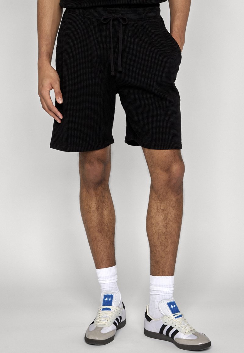 Homme portant un short noir texturé à cordon de serrage, des chaussettes blanches et des baskets blanches avec des rayures noires et des patchs étoile bleue.