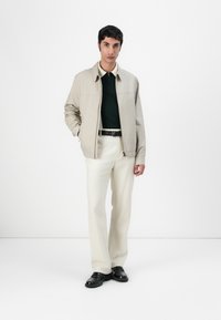 Lichtbeige jack met een ritssluiting, gedragen over een donkergroen poloshirt. Crèmekleurige broek en zwarte schoenen maken de outfit compleet.