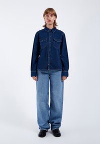 Dunkelblauses Denim-Hemd mit Druckknopfleiste, zwei Brusttaschen und Kragen. Kombiniert mit hellblauen weit geschnittenen Jeans und schwarzen Schuhen.