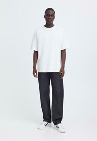 T-shirt bianca oversize, con maniche larghe, abbinata a jeans dritti blu scuro e sneakers bianche con strisce nere. Sfondo neutro.