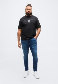 Άνδρας με πλεξούδες που φοράει μαύρο t-shirt Tommy Hilfiger, μπλε τζιν και λευκά αθλητικά παπούτσια, στέκεται με το ένα χέρι στην τσέπη.