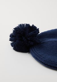 Marineblå strikket vinterlue med en stor pom-pom på toppen, vist mot en enkel lys bakgrunn.