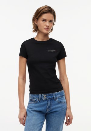 Mladá žena s krátkymi vlasmi, oblečená v priliehavom čiernom tričku „tommy jeans“ a modrých džínsových nohaviciach s vysokým pásom na bielom pozadí.