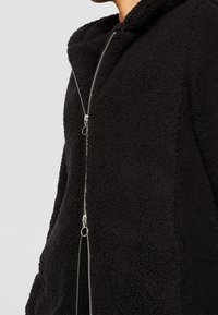 Chaqueta de forro polar negra con capucha, que presenta un cierre frontal de cremallera completa, una tela texturizada y herrajes de cremallera en tono plateado.
