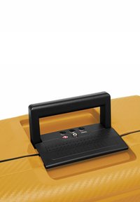 Il manico della valigia gialla presenta una presa nera testurizzata e aperture per lucchetti, con un design elegante e una finitura liscia.