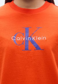 Πορτοκαλί βαμβακερό μπλουζάκι με μεγάλο μπλε "CK" και "CalvinKlein" σε λευκό. Στρογγυλή λαιμόκοψη και κοντά μανίκια με λείο ύφασμα.