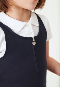 Rochie bleu marin cu un fermoar și un prindere în formă de inimă, purtată peste o cămașă albă cu guler. Rochia are o textură netedă și un design ajustat.