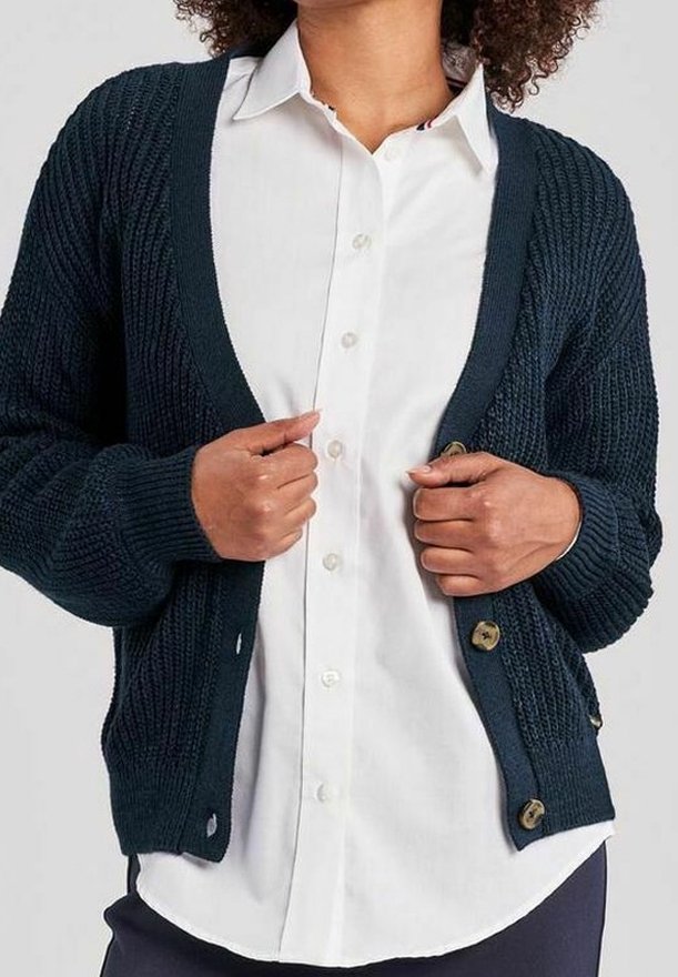 Henkilö, jolla on yllä valkoinen nappikauluspaita ja tumma sininen neulecardigan, pitämällä cardiganin reunoista nappien lähellä.