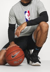 Camiseta gris con el logo de la NBA sobre una camiseta de manga larga negra, pantalones cortos negros, zapatos Nike negros, sosteniendo un balón de baloncesto marrón con las marcas Nike y Elite.