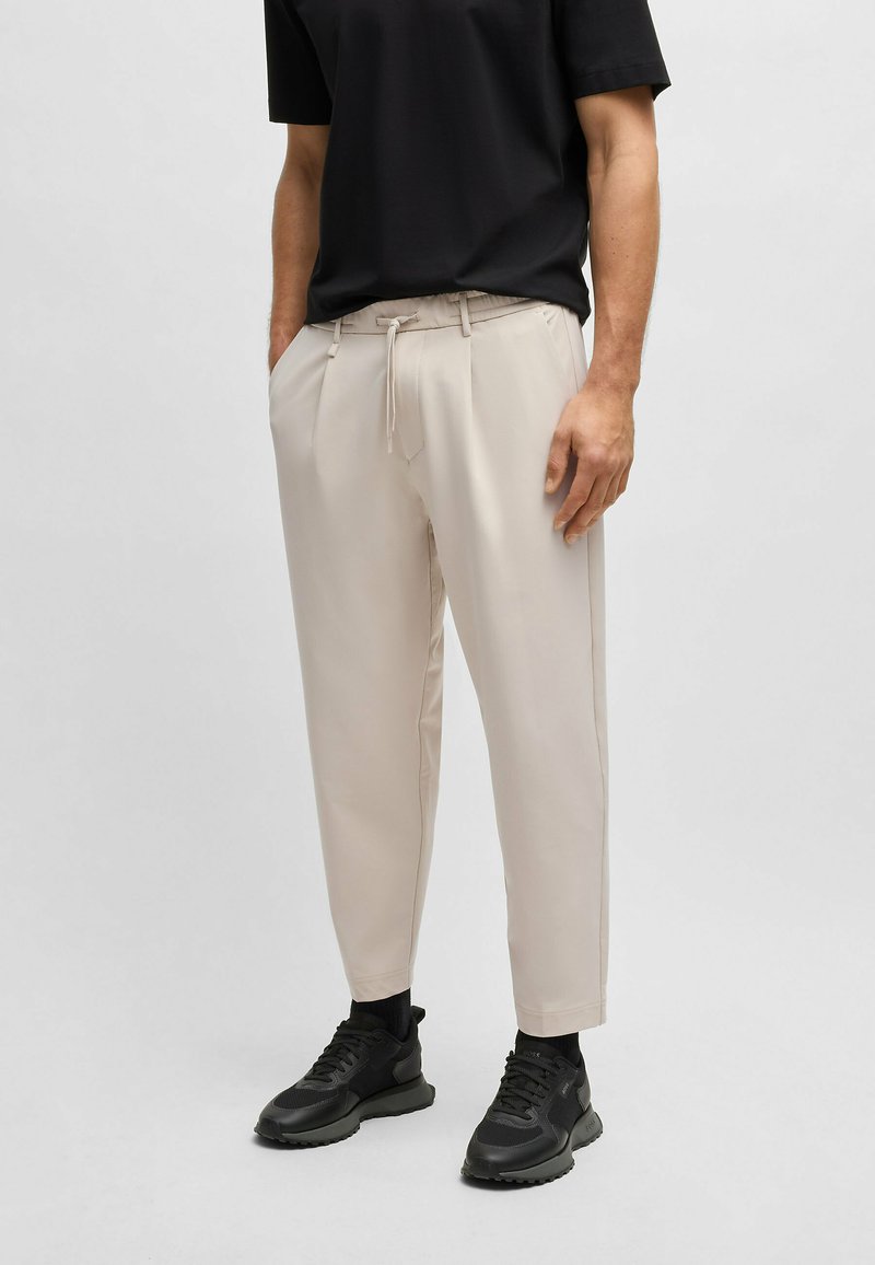 Beige taperende broek met voorplooien en zijzakken, met een elastische tailleband en trekkoord. Gemonteerd met zwarte sneakers.