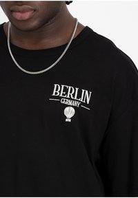 T-shirt en coton noir avec texte brodé blanc "BERLIN ALLEMAGNE" et graphique de ballon à air chaud, col rond et coupe décontractée. Collier en chaîne argenté.