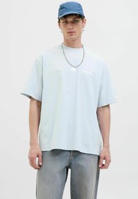 Helles blaues Oversize-T-Shirt aus Baumwolle, mit kurzen Ärmeln und einem dezenten Logo. Begleitet von verwaschenen Jeans und einer blauen Kappe.