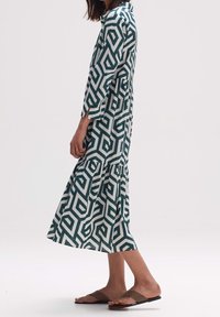 Lång klänning i grönt och vitt geometriskt mönster, med krage, trekvartslånga ärmar och ett flerlagerskjol. Slät tygtextur.