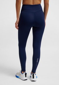 Granatowe legginsy z wysokim stanem, wykonane z elastycznego materiału; charakteryzują się gładką teksturą, dopasowanym krojem oraz logo blisko kostki.