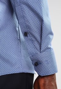 Blauw overhemd met knopen, voorzien van een rond patroon op een gestructureerde stof. Opvallende contrasterende manchetten en donkere knopen.