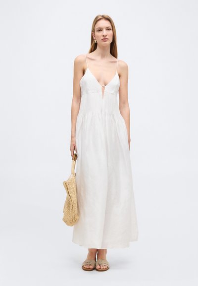 Žena v bielych maxi šatách bez rukávov s tenkými ramienkami, držiaca tkanú kabelku a obutá v tkaných nazouvacích sandáloch na jednoduchom pozadí.