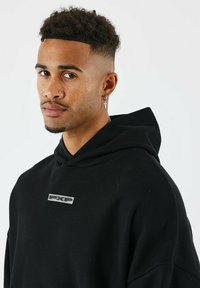 Sudadera negra con un acabado texturizado, que presenta un logo plateado en el pecho y una capucha amplia. El modelo tiene el cabello corto y rizado, además de pendientes.