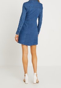 Džínové šaty s dlouhými rukávy, přiléhavým pasem a rozšířeným spodním okrajem. Světle modrá barva s prošíváním; nošeny s bílými sandály na podpatku.
