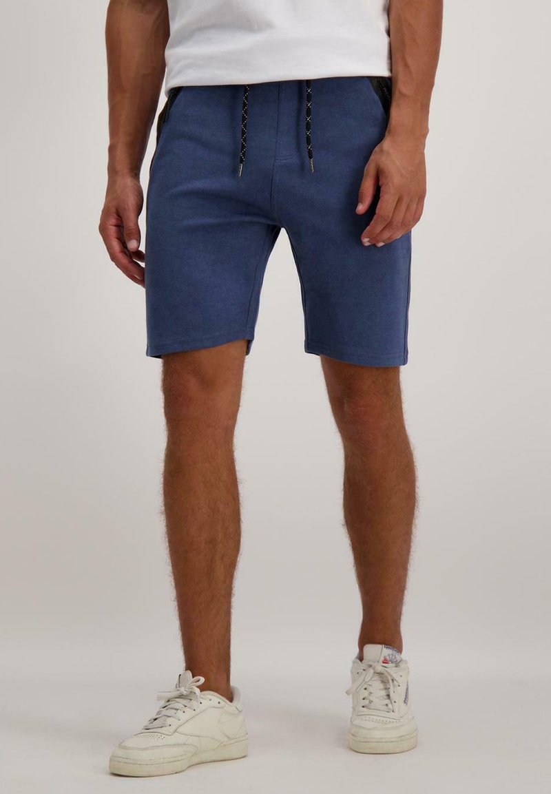 Blauwe katoenen shorts met een elastische tailleband, platte koordsluiting en zijzakken. Gemaakt met witte sneakers op een neutrale achtergrond.