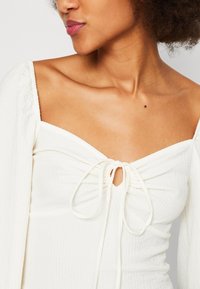Frau mit natürlichem lockigem Haar, die ein weißes, strukturiertes Off-Shoulder-Oberteil mit einer Front-Schleife trägt.