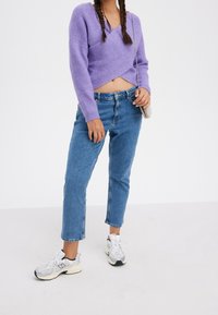 Lawendowy sweter z dzianiny o fasonie chusty, wysokiej talii niebieskie dżinsy oraz białe trampki ze srebrnymi akcentami, noszone z torebką na łańcuchu na ramię.