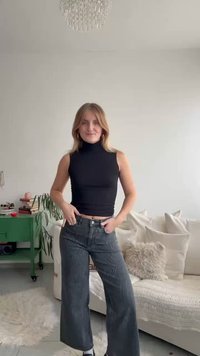 Top preto sem mangas com gola alta combinado com jeans escuros de perna larga com padrão subtil, usado com ténis brancos.