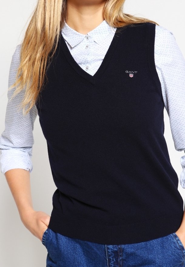 Gilet en tricot bleu marine avec un col en V, ourlet côtelé et petit logo GANT. Porté par-dessus une chemise à boutons à motifs bleu clair.