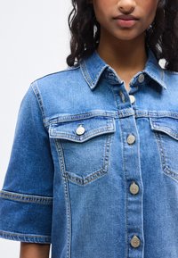 Πουκάμισο από τζιν σε ανοιχτό μπλε χρώμα με κοντά μανίκια, εμπρόσθιο κούμπωμα με κουμπιά, δύο τσέπες στο στήθος και μεταλλικά κουμπιά. Διαθέτει κοντράστ ραφές και κλασικό γιακά.