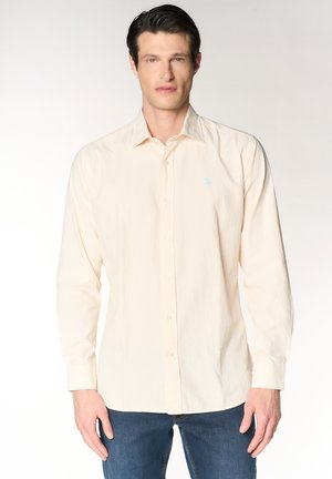 Uomo che indossa una camicia beige chiaro a righe con bottoni e maniche lunghe e jeans blu, in piedi davanti a uno sfondo bianco semplice.