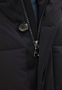 Svart puffjacka med dragkedjestängning, glänsande knapp och textil med struktur. Har en ribbad krage och kontrasterande foder synligt inuti.