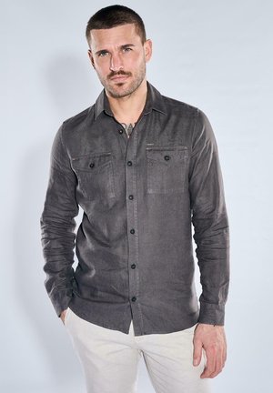 Mann mit kurzen Haaren und Bart, der ein dunkelgraues Hemd mit Knöpfen und hellfarbene Hose trägt, steht mit einer Hand in der Tasche.
