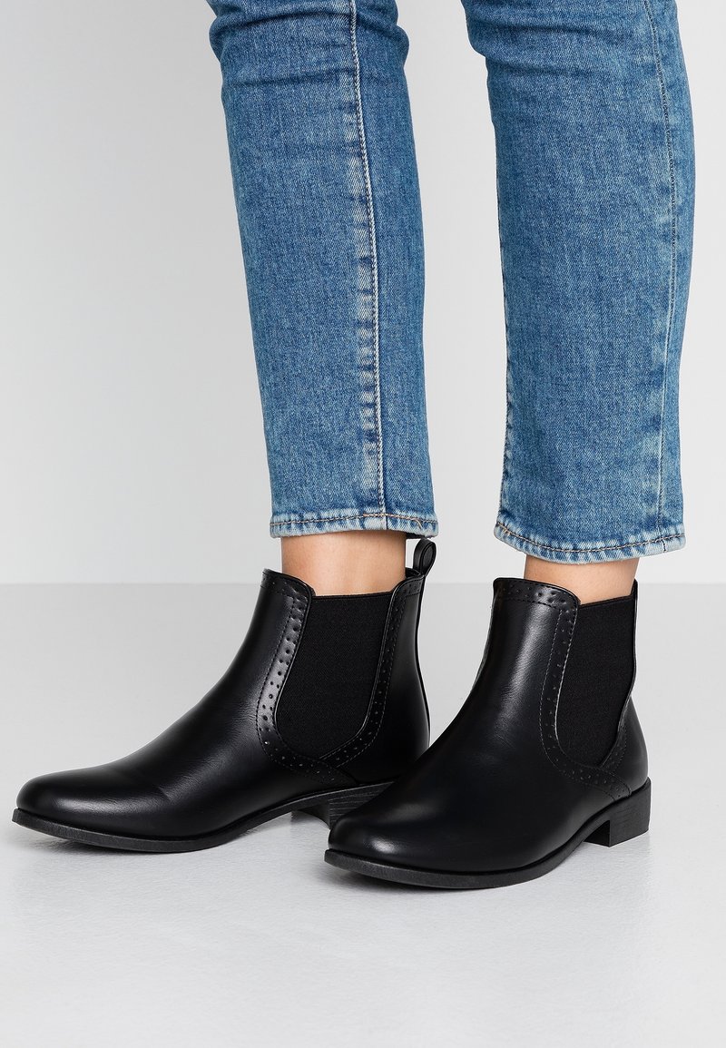 Bottines en cuir noir avec panneaux latéraux élastiques portées avec un jean bleu retroussé sur un fond de sol et de mur clair.