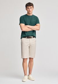 Camiseta de algodón verde con un pequeño logotipo, pantalones cortos beige, cinturón marrón y zapatillas blancas. Ajuste casual, textura suave, mangas cortas.