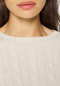 Světlý béžový pletený svetr se širokým žebrovaným výstřihem a copánkovým vzorem, který nabízí texturovaný a měkký vzhled.