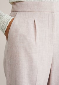 Ruka u džepu visokog struka, svijetloružičastih hrapavih hlača uparena s dugom bijelom rebrastom majicom s dugim rukavima.