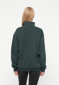 Σκούρο πράσινο πουλόβερ με υψηλό λαιμό, ριμπ μανσέτες και χαλαρή εφαρμογή. Μαλακή υφή, μίνιμαλ σχέδιο, με μια μικρή ετικέτα στο πίσω μέρος.