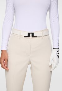 Housut beige, jotka on räätälöity ja joissa on sileä pinta, varustettuna valkoisella vyöllä ja mustalla neliönmuotoisella soljella. Alla valkoinen pitkähiha. Kädessä valkoinen golf-käsine.