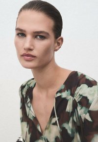 Blusa ligera con estampado, escote en V profundo y mangas fruncidas. Los colores incluyen verde, marrón y crema, con una textura de tejido suave.