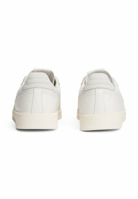 Zapatillas de cuero blancas con textura suave, cuello acolchado y suela de goma. Presentan un diseño minimalista y sutiles detalles de costura en la parte posterior.