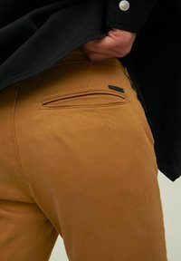 Brown cotton trousers with a back pocket featuring a small black label, and a slightly textured surface, paired with a black garment.