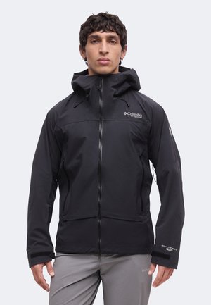 Mann trägt schwarze Columbia Titanium wasserdichte Jacke mit Kapuze und graue Hose, steht vor einfarbigem Hintergrund.