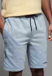Shorts en polaire bleu clair avec une taille élastique, un cordon de serrage et des accents imprimés sur les côtés. Présente une surface texturée et douce.