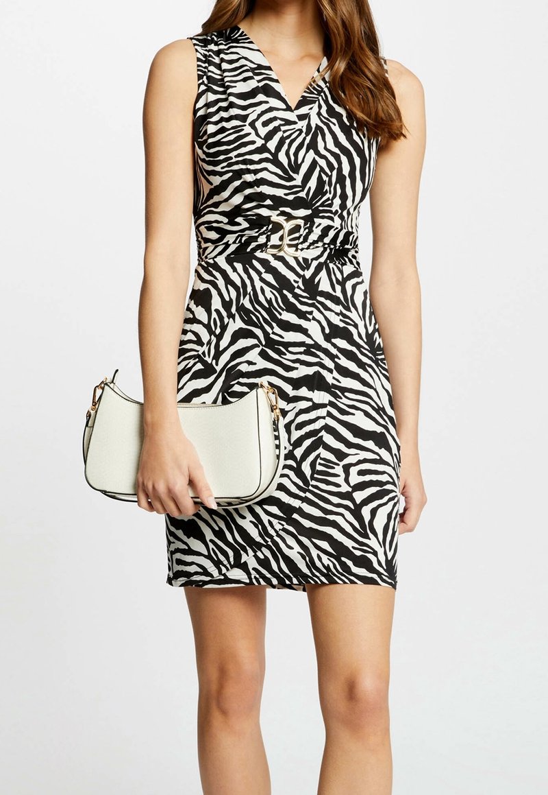 Schwarz-weißes Kleid mit Zebramuster, V-Ausschnitt und tailliertem Gürtel. Hält eine kleine weiße Tasche mit geschwungener Form und goldenen Metallakzenten.