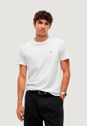 Giovane uomo con capelli ricci che indossa una t-shirt bianca, pantaloni neri, cintura nera e orologio da polso, in piedi davanti a uno sfondo chiaro e uniforme.