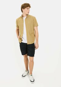 Korte mouwen, geknoopte shirt in geel en bruine ruitpatroon, gecombineerd met zwarte shorts en witte sneakers. Zichtbare knopen en zakken.